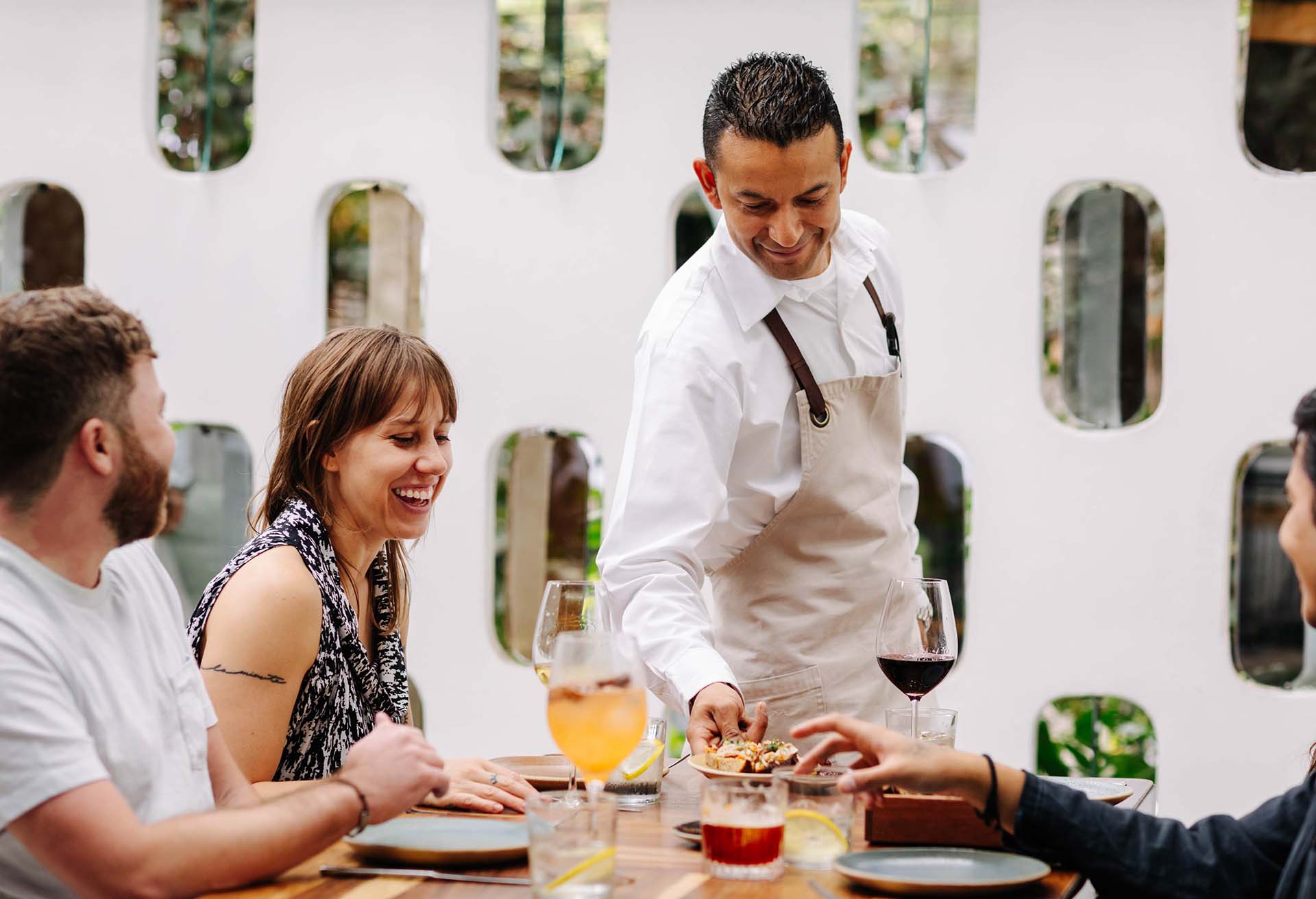 Image depicts a restaurant scene where a waiter is serving dishes at a table. Guests are smiling and appear entertained