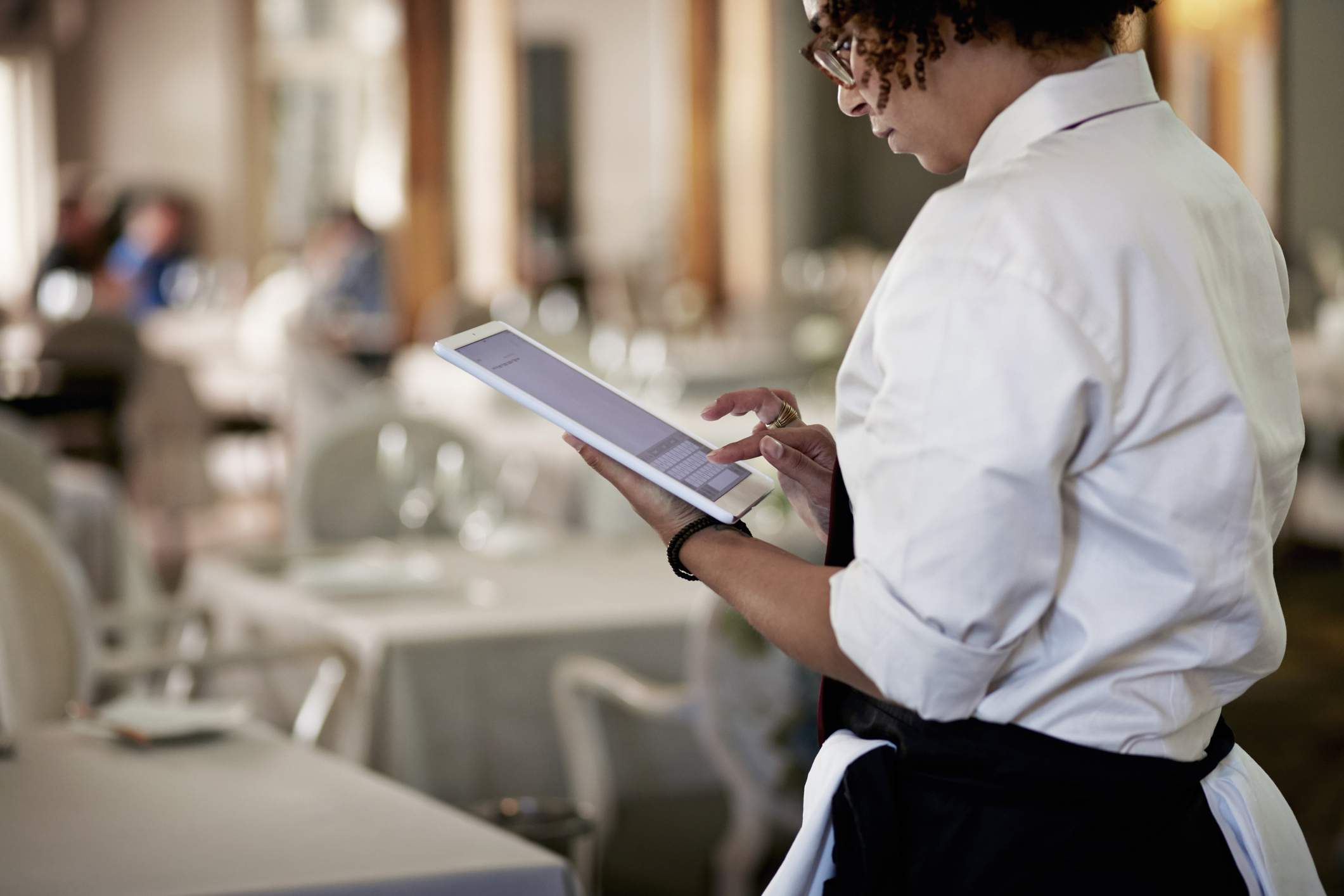 Image depicts a server wearing a white button down and black apron using a tablet inside of a restaurant. 
