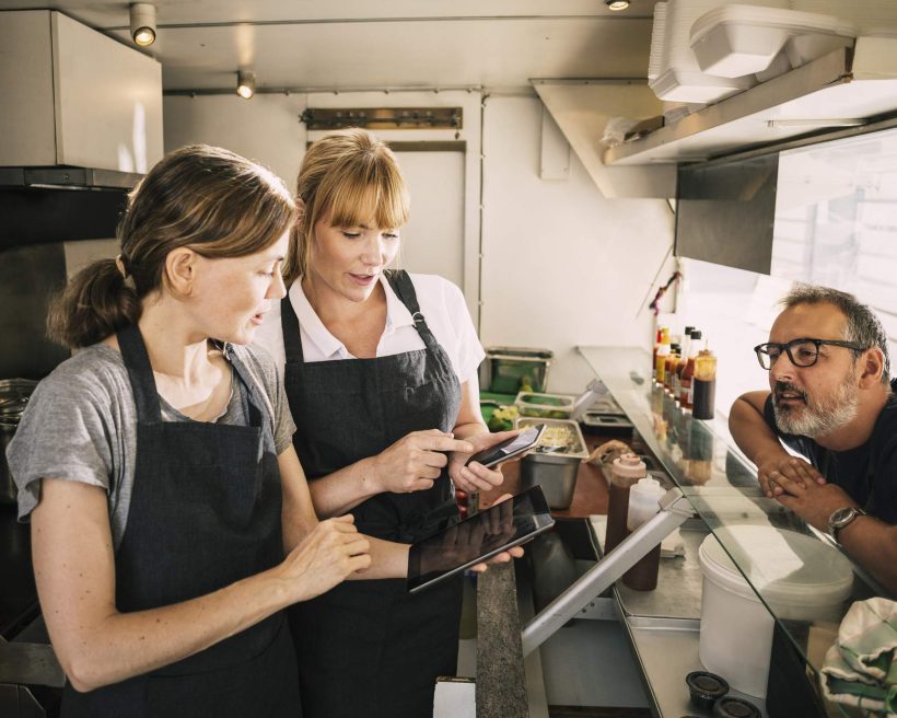 Three restaurant employees are having a conversation. One of them is holding a mobile phone and and showing some content to the other employee.