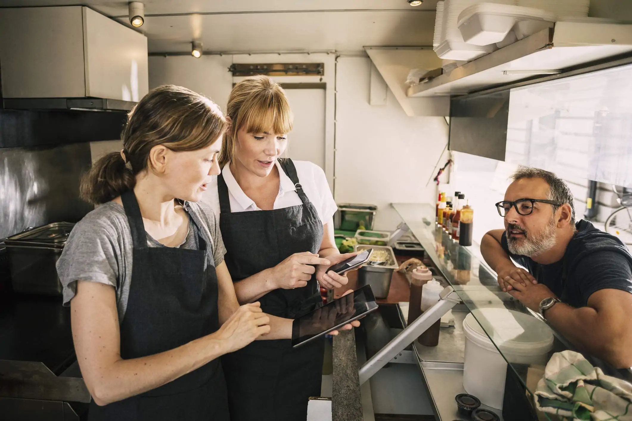 Three restaurant employees are having a conversation. One of them is holding a mobile phone and and showing some content to the other employee.