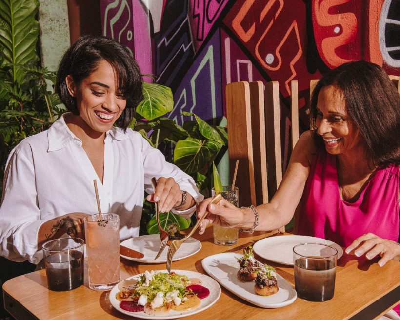 Two women dining at a restaurant with a colourful wall in the background and a table showcasing an array of dishes and drinks